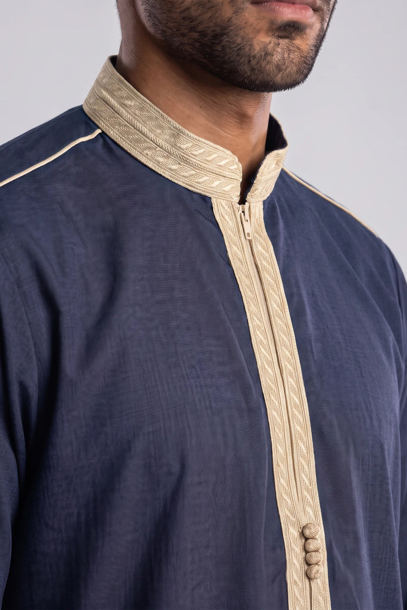 A man modeling a long-sleeved navy blue traditional robe with beige trim and a beige zipper placket, standing against a neutral gray studio background.