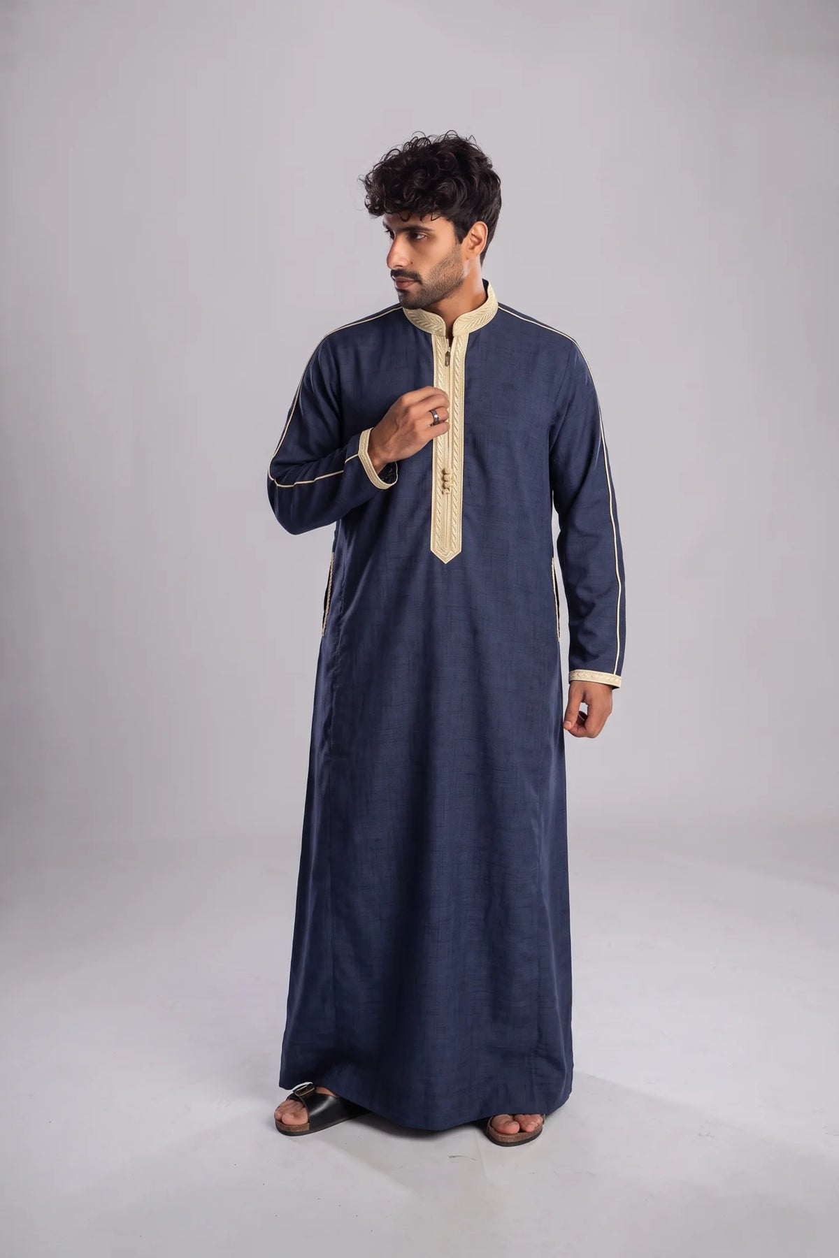 A man modeling a long-sleeved navy blue traditional robe with beige trim and a beige zipper placket, standing against a neutral gray studio background.