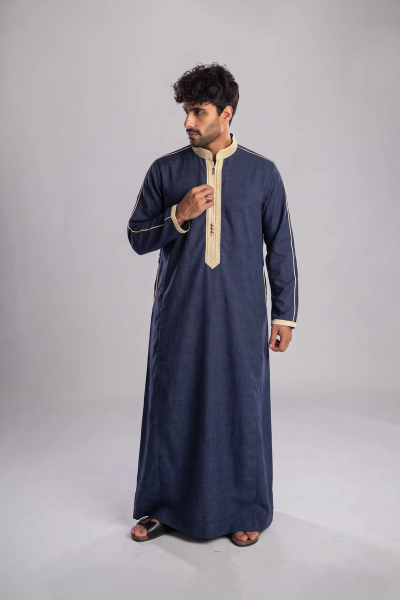 A man modeling a long-sleeved navy blue traditional robe with beige trim and a beige zipper placket, standing against a neutral gray studio background.